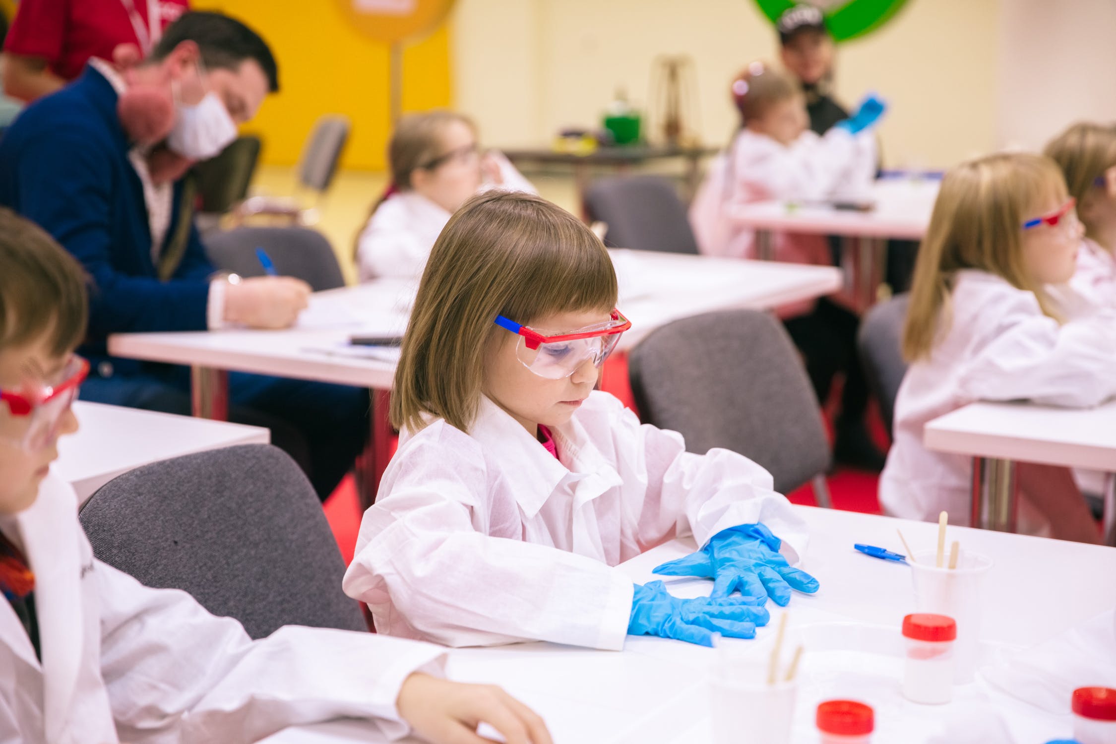 young children sit at desks, dressed in lab gear