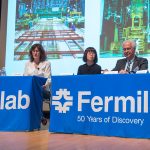 Four people sitting on a panel on a stage with a Fermilab tablecloth draped over the table.