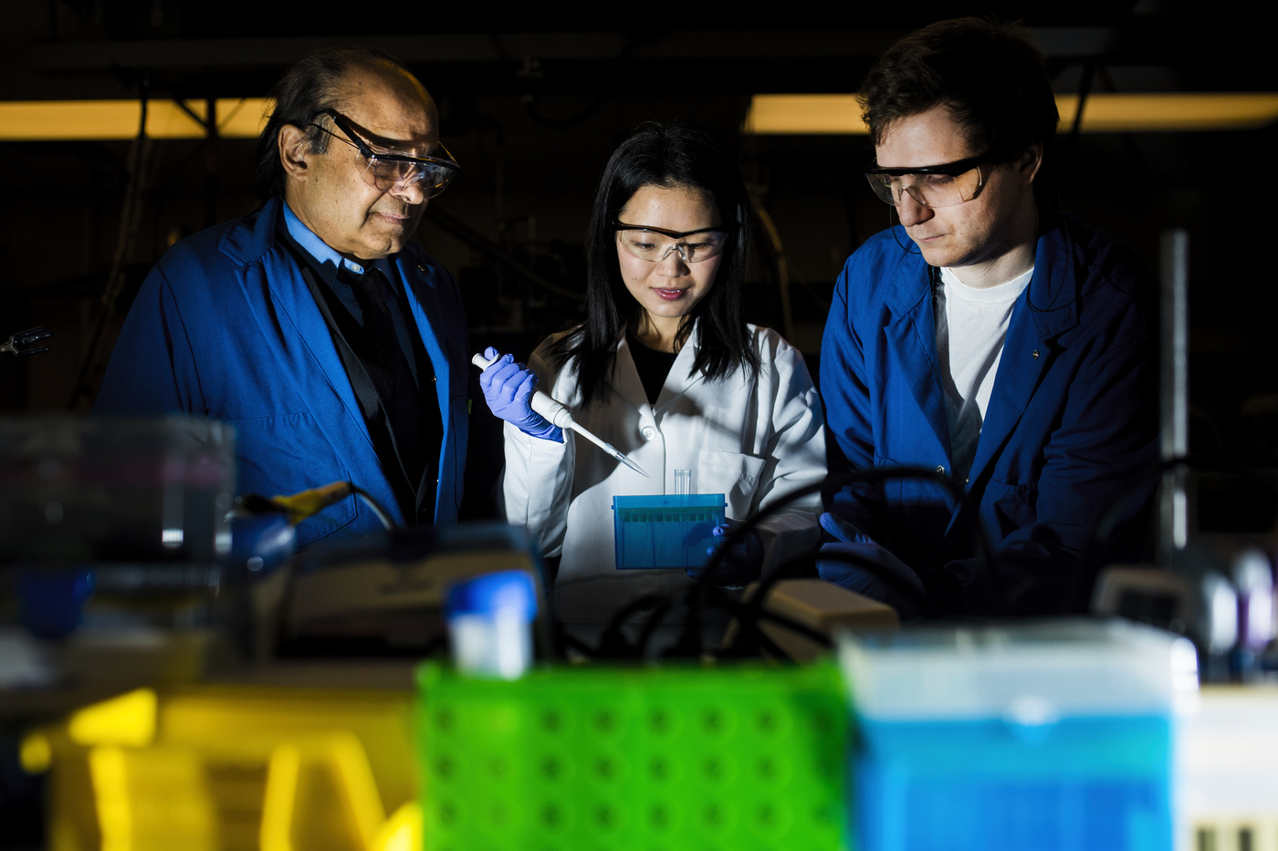 Ju Qiao is holding a pipette while flanked by Sri Sridhar and Liam Timms in a darkened lab.