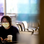 a student sits at a desk, masked up, doing work on her laptop