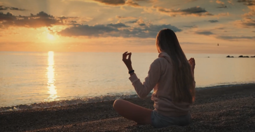the back of a girl facing the ocean, sitting criss cross on the shore, meditating as the sun sets