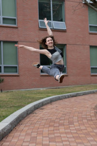 katherine leaps in the air and poses on a brick walkway with a brick building behind her