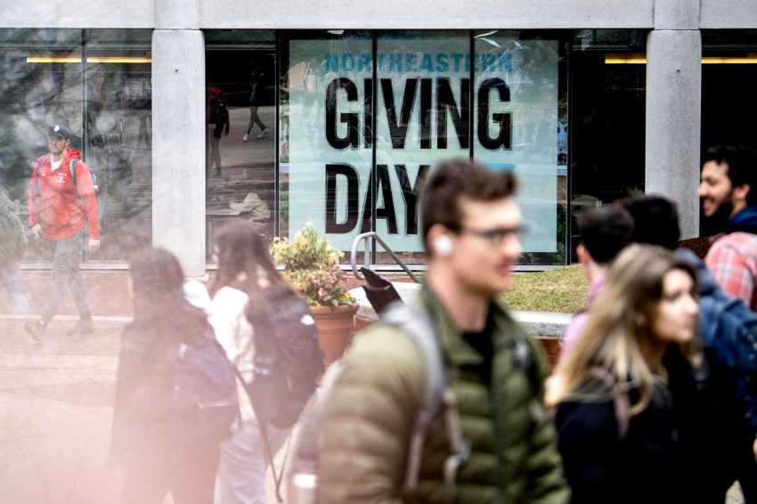 students walking across campus in front of a building with a giving day poster in the window