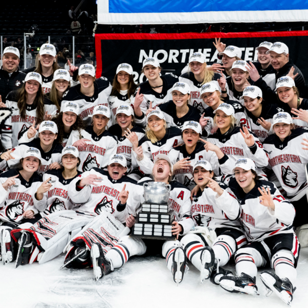 Photo of a women's hockey team sitting on the ice with a trophy. Behind them is the edge of a blue and white banner with a black and red bottom. There is a husky mascot sitting with the team as well.