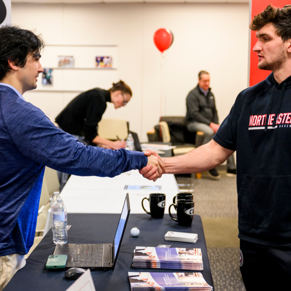 Two men shake hands over a table. Table has papers and various things on it. The man on the left is wearing khakis and a blue shirt. Man on the right is wearing a black Northeastern Athletics top and carrying a blue water bottle.