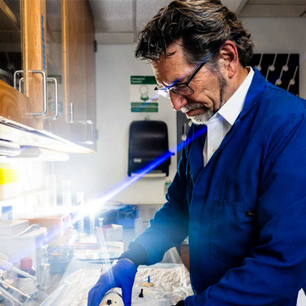 Man in a blue lab coat and blue latex gloves stands with his hands on lab equipment in a lab. He is wearing safety glasses.