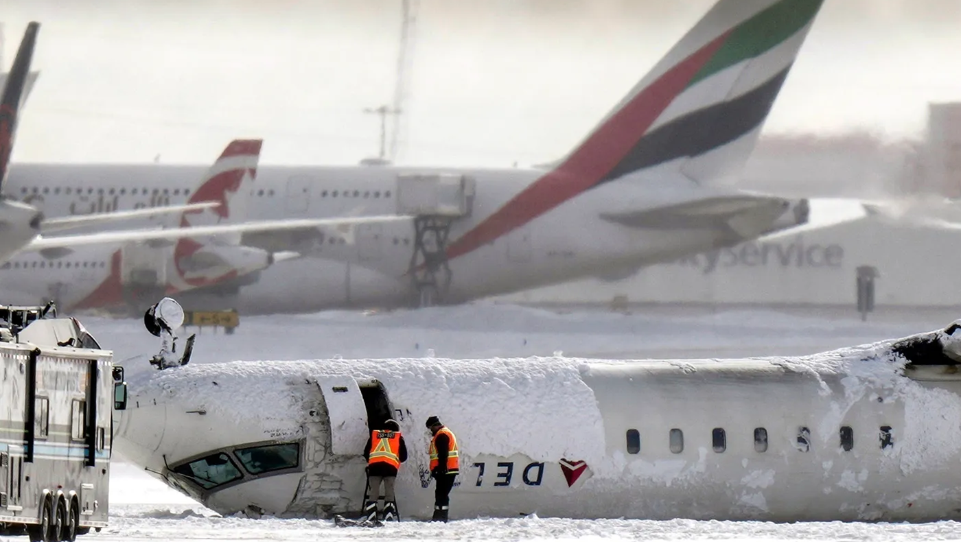 Delta plane upside down on a runway as two people wearing orange vests stand in front of it, looking into the plane. There is snow on the ground and other planes in the background.