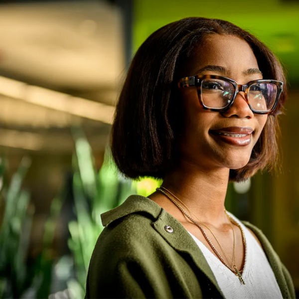 Young black woman with glasses in an open green shirt with a white shirt underneath. Background is blurred with green and brown