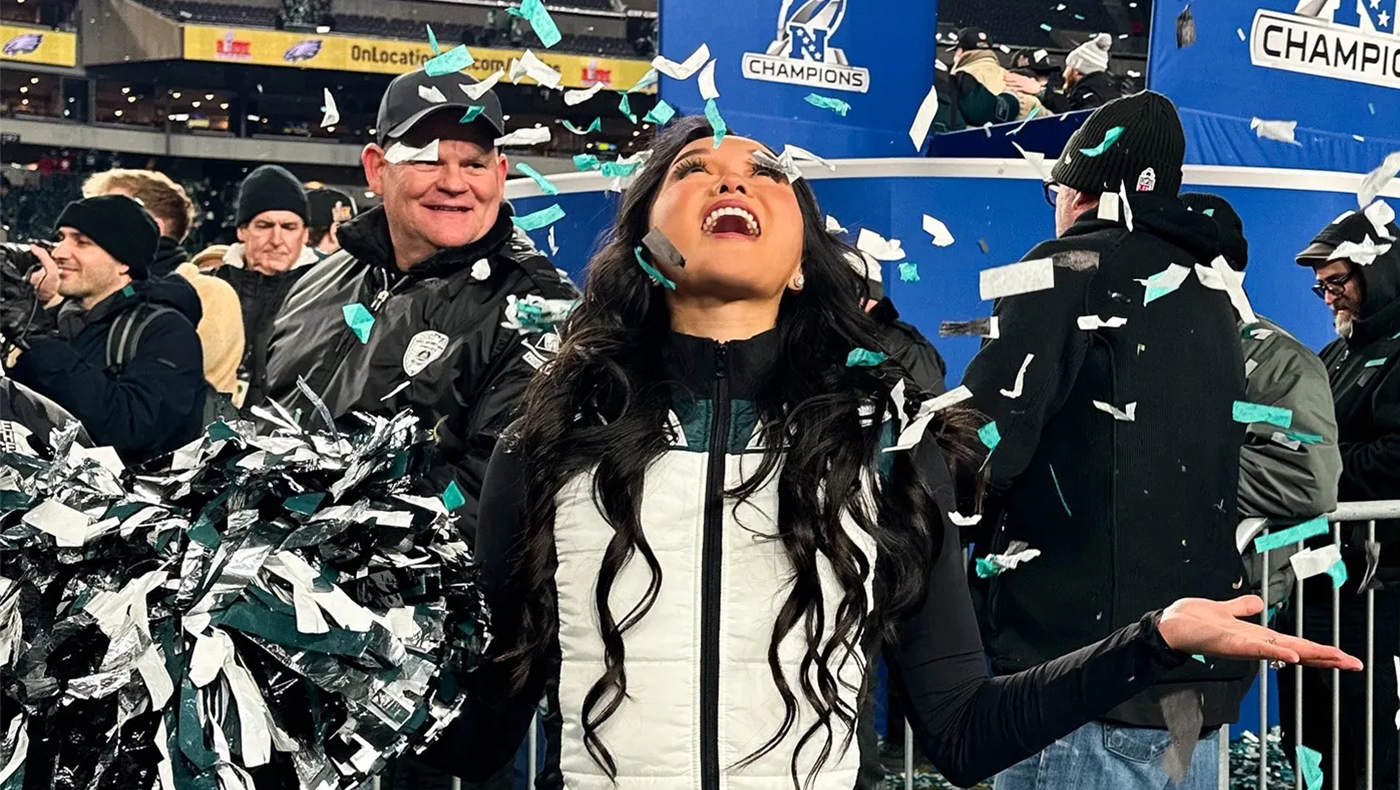 Women in a white and black jacket stands with her face and hands upturned, as confetti falls around her. She is holding a green and white pom pom in her right hand.