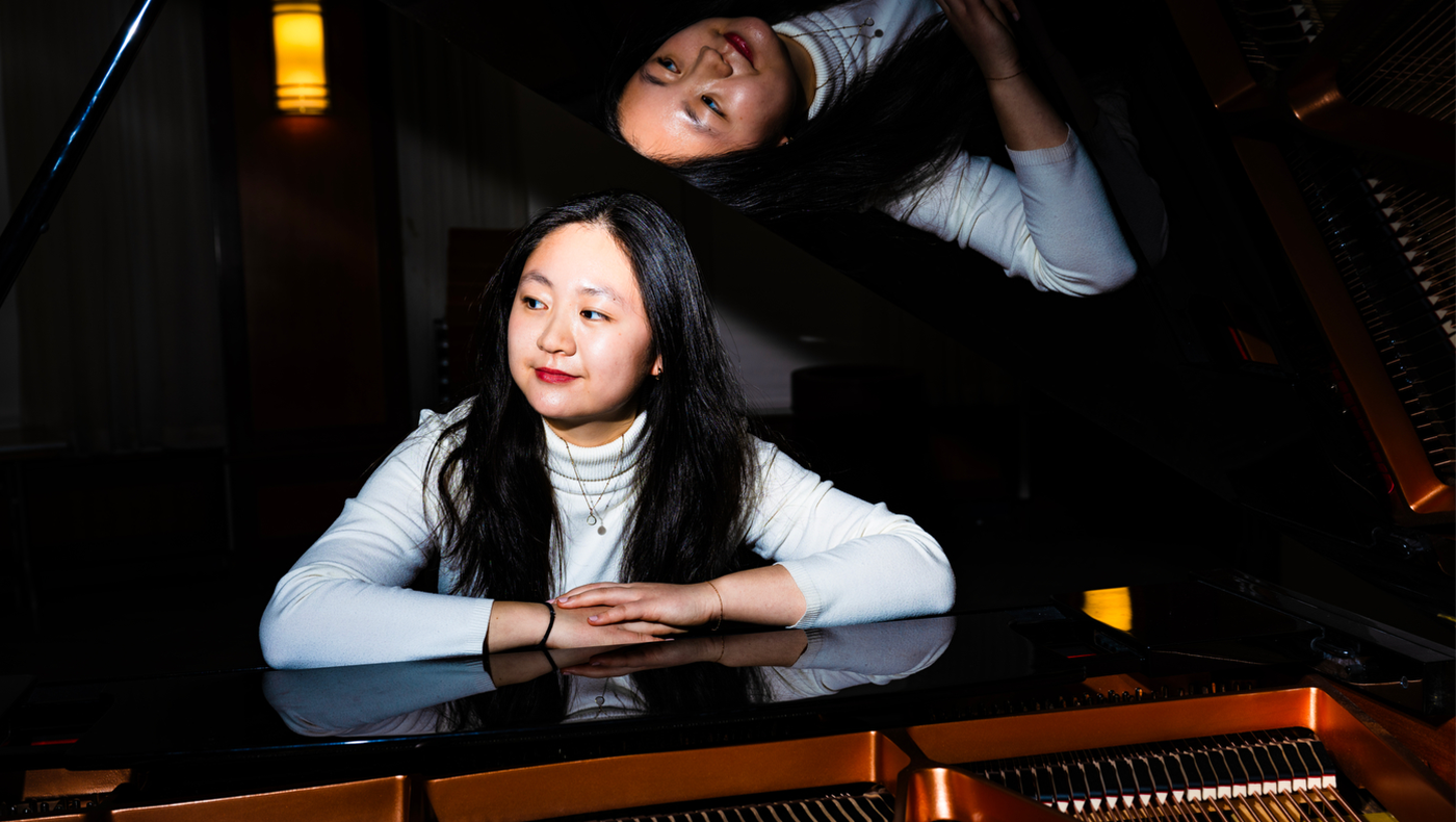 Young woman in a white turtle neck sweater sits at a piano with her hands folded on top of it. She is looking off to the left.