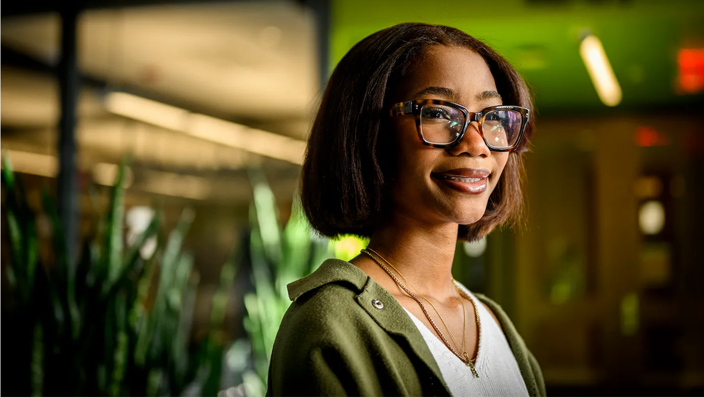 Young black woman with glasses in an open green shirt with a white shirt underneath. Background is blurred with green and brown
