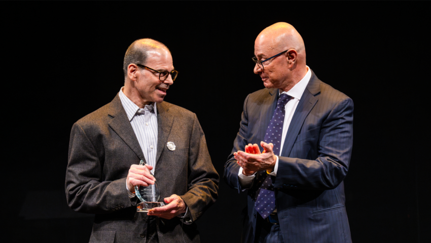 Two men in suits in front of a black background. The man on the right is clapping and looking to the left. The man on the left is holding a glass award and looking towards the right.