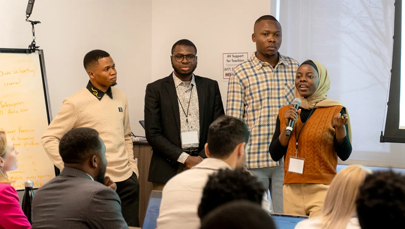 Photo of four people speaking to a room of others. The group consists of 3 men and one woman all dressed in semi professional clothing. The woman is holding a mic and adressing the crowd.