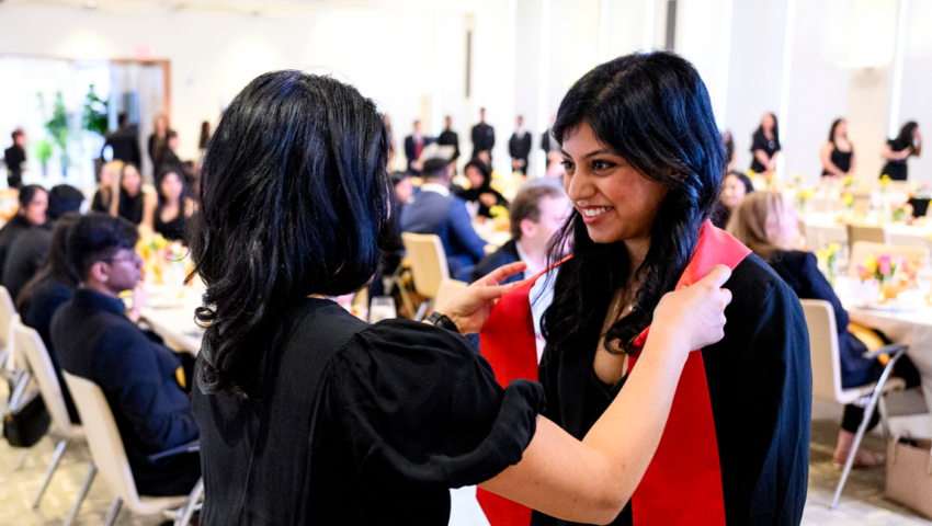 Photo if a woman with black hair laying a stole over a young woman's head. Both women are wearing black and the sole is red. In the background are people sitting at banquet tables.
