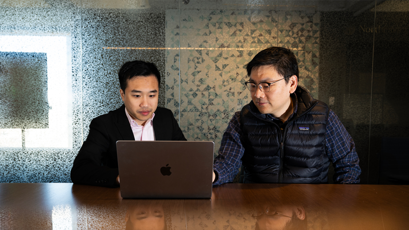 Two men sit at a table looking at an apple laptop.