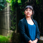 Women in a blue shirt and a black blazer stands in a garden. Her hands are clasped in front of her and she is looking off to the left.