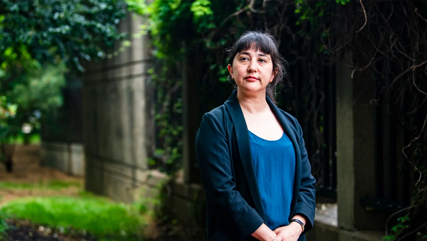 Women in a blue shirt and a black blazer stands in a garden. Her hands are clasped in front of her and she is looking off to the left.