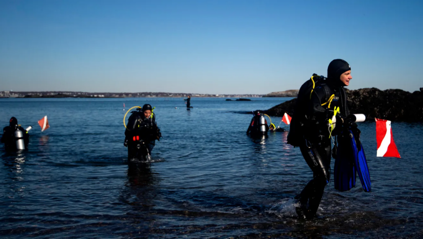 Photo of a scuba diver getting out of the Ocean. The diver is wearing a wet suit and carrying a red and white flag. Behind him there are other divers exiting the water.
