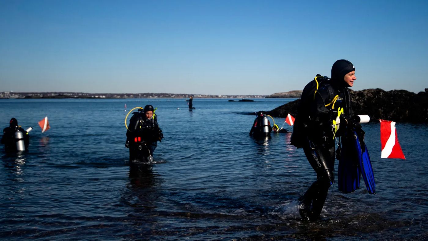 Photo of a scuba diver getting out of the Ocean. The diver is wearing a wet suit and carrying a red and white flag. Behind him there are other divers exiting the water.