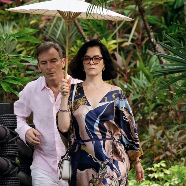 Screen shot of a scene from Season 3 of The White Lotus. Features a woman in a blue patterned dress carrying an umbrella while a man in a pink button down hurries after her. They are surrounded by greenery.