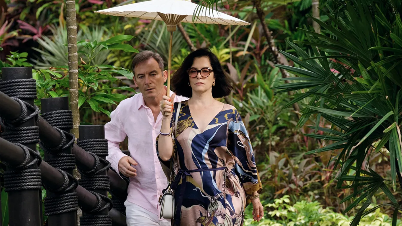 Screen shot of a scene from Season 3 of The White Lotus. Features a woman in a blue patterned dress carrying an umbrella while a man in a pink button down hurries after her. They are surrounded by greenery.