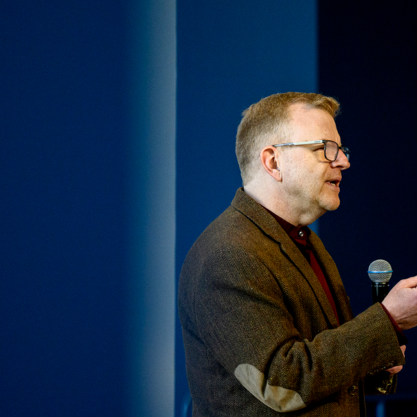 Picture of a man in a black suit jacket and button down talking into a microphone. He is wearing glasses and turned to the left. The background is blue.